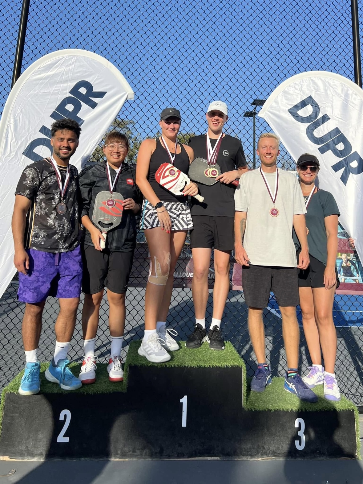 Sueme Rajpal displaying silver medal and trophy at a pickleball tournament in Australia with partner Dora Tsent, while standing on a podium with other professional pickleball players Katy Hart, Ollie Gray.